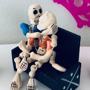 May include: A handcrafted Day of the Dead figurine depicts two skeleton figures embracing on a black bench. The skeletons are painted with off-white, orange, and blue accents. The bench has a decorative white cross design.