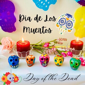 May include: A vibrant Day of the Dead display featuring colorful sugar skulls, lit red candles, and floral accents. The text "Dia de Los Muertos" and "Day of the Dead" are displayed in elegant script.