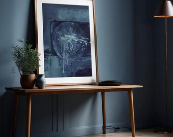 Puede incluir: Una impresión artística abstracta enmarcada, en tonos azules y blancos, se apoya contra una pared azul oscuro. La obra de arte se exhibe sobre una mesa larga de madera, junto a una planta en maceta y un pequeño jarrón. Una lámpara de pie con pantalla oscura también es visible.