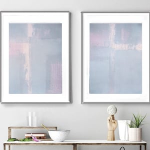 Könnte beinhalten: Zwei gerahmte abstrakte Gemälde mit hellblauem Hintergrund und weißen und rosafarbenen Pinselstrichen. Die Gemälde hängen an einer weißen Wand.