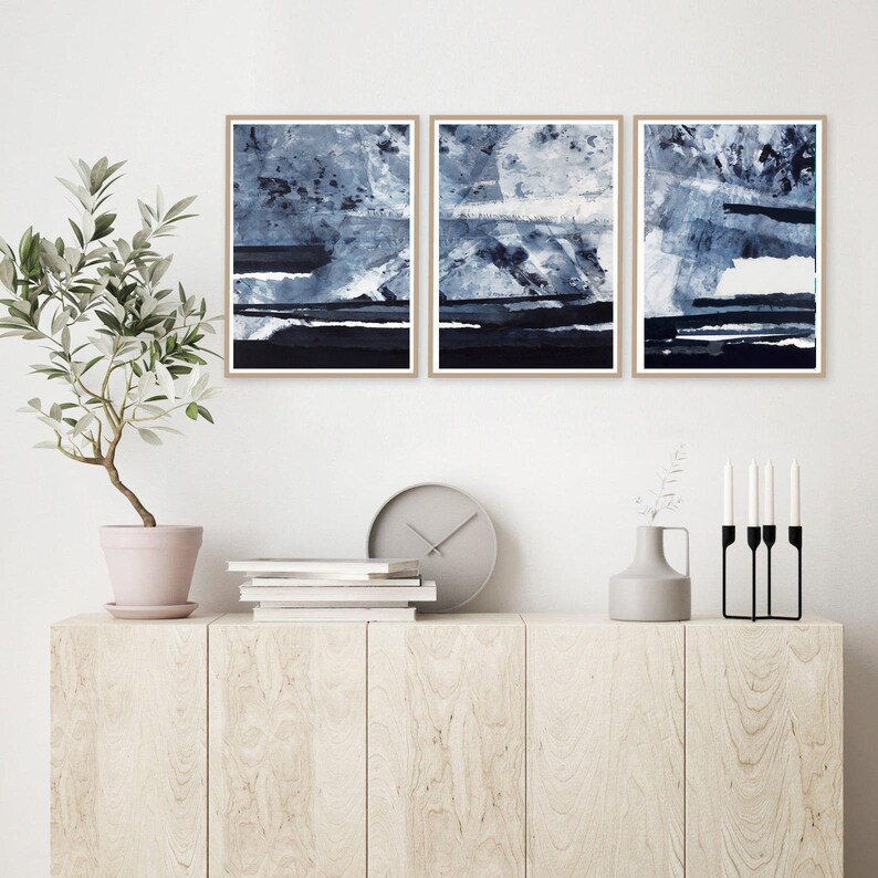 Pu&ograve; includere: Tre dipinti astratti in tonalit&agrave; di blu e bianco, incorniciati in legno naturale, appesi a una parete bianca sopra un mobile in legno con una pianta in vaso, libri, un orologio e un vaso.
