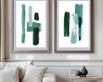 May include: Two framed abstract art prints. Each print features brushstrokes in shades of teal and dark green on a white background. The frames are a muted silver color. The artwork is displayed above a white sofa.