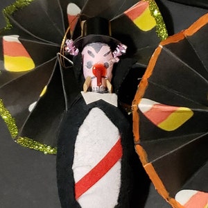May include: A black and white Halloween decoration of a vampire with a red nose and fangs. The vampire is wearing a black top hat and a white shirt with a red stripe. The decoration is surrounded by two black paper fans with candy corn designs.
