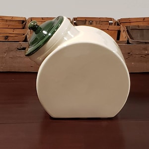 May include: Cream-colored ceramic canister with a dark green lid. The canister has a rounded rectangular shape and a tilted lid. The lid has a small knob. The background features wooden baskets.