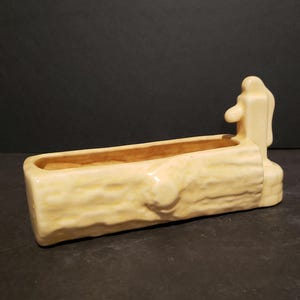 May include: A ceramic planter shaped like a log with a small spout on the side. The planter is a light yellow color and has a rough, textured surface.