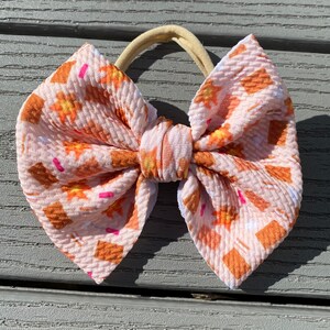 May include: A pink and orange patterned hair bow with a brown elastic headband. The bow is made of a soft, textured fabric.
