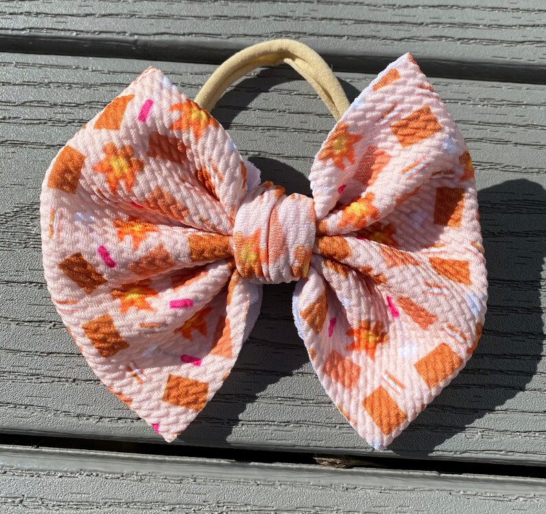 May include: A pink and orange patterned hair bow with a brown elastic headband. The bow is made of a soft, textured fabric.