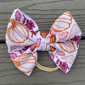 May include: A white fabric bow with a pumpkin and leaf pattern in shades of orange, brown, and pink. The bow is tied with a brown elastic headband.