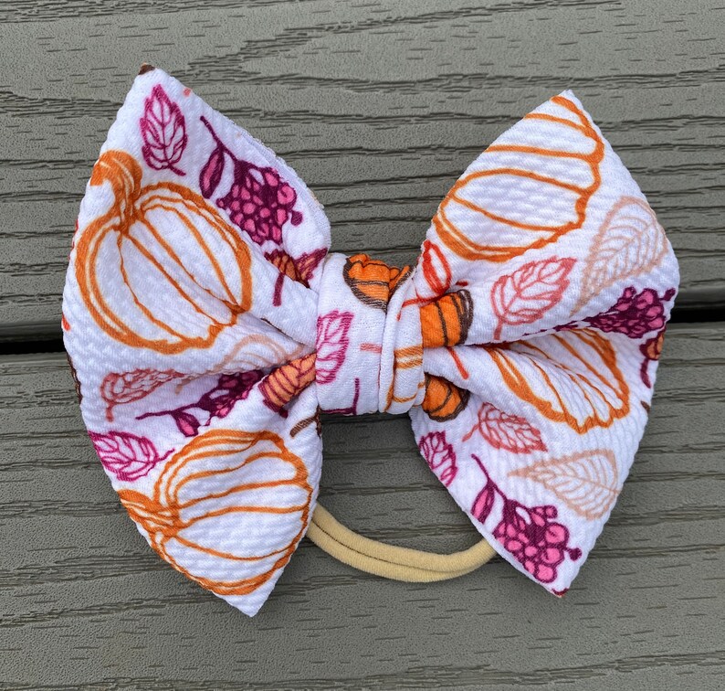 May include: A white fabric bow with a pumpkin and leaf pattern in shades of orange, brown, and pink. The bow is tied with a brown elastic headband.