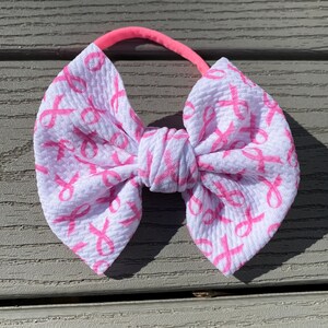 May include: A white headband with a pink ribbon bow. The bow is made of fabric with a pink ribbon pattern.