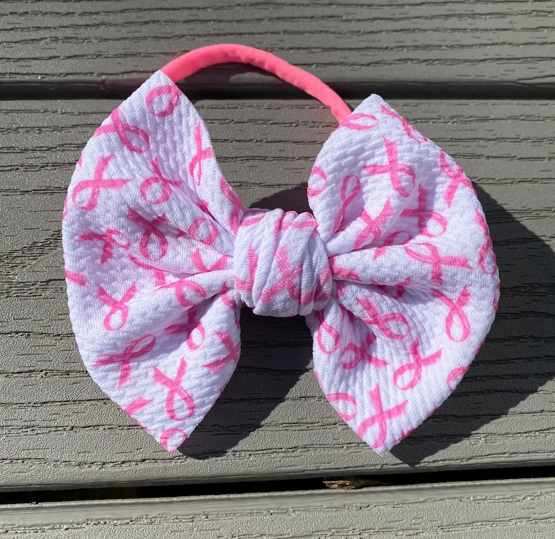 May include: A white headband with a pink ribbon bow. The bow is made of fabric with a pink ribbon pattern.