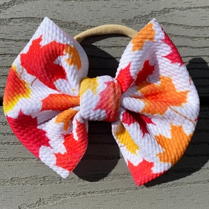 May include: A white headband with a large bow made of fabric with a red, orange, and yellow leaf print.
