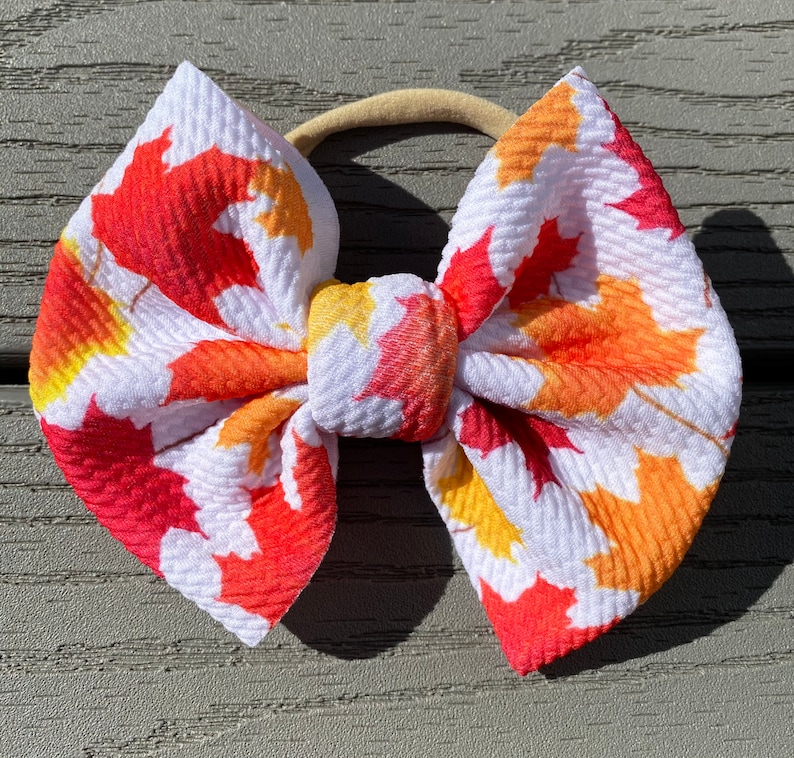 May include: A white headband with a large bow made of fabric with a red, orange, and yellow leaf print.