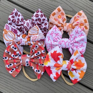 May include: Six different patterned hair bows on nude elastic headbands. The patterns include footballs, pumpkins, pink ribbons, owls, and fall leaves.