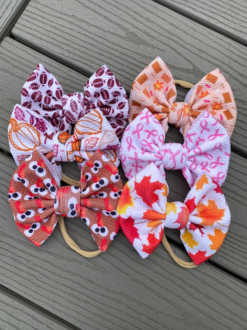 May include: Six different patterned hair bows on nude elastic headbands. The patterns include footballs, pumpkins, pink ribbons, owls, and fall leaves.