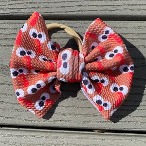 May include: A brown and red bow with a turkey face pattern. The bow is tied with a nude elastic headband.