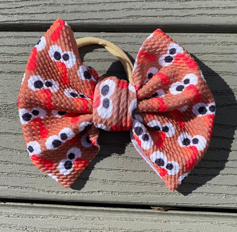 May include: A brown and red bow with a turkey face pattern. The bow is tied with a nude elastic headband.