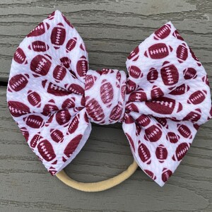 May include: White headband with a brown football pattern bow. The headband is made of elastic and is suitable for babies and toddlers.