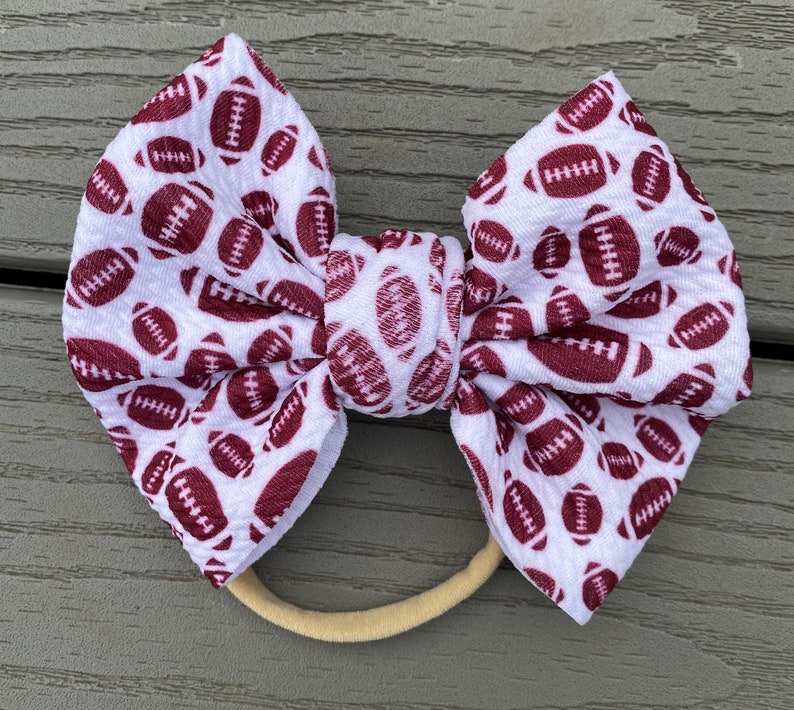 May include: White headband with a brown football pattern bow. The headband is made of elastic and is suitable for babies and toddlers.