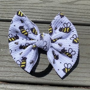 May include: A white hair bow with a black elastic band. The bow is covered in a white fabric with a repeating pattern of black and yellow bees.