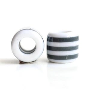 Puede incluir: Dos cuentas de cerámica blanca con un agujero en el centro. Una cuenta tiene forma de anillo, la otra es cilíndrica con rayas grises horizontales. Las cuentas están sobre un fondo blanco.