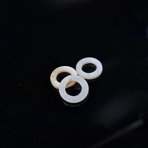 May include: Three small, round, mother-of-pearl beads with a hole in the center. The beads are off-white and iridescent, with a slight variation in color. They are arranged on a black surface.