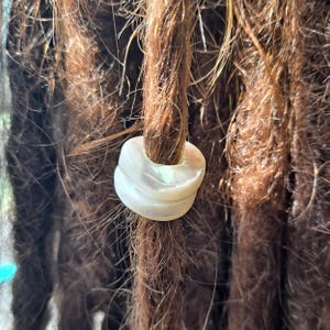 Könnte beinhalten: Nahaufnahme von braunen Dreadlocks mit einer weißen, irisierenden Haarperle. Die Perle ist kreisförmig und hat ein geschichtetes Design. Das Haar ist dunkelbraun und hat eine natürliche Textur.