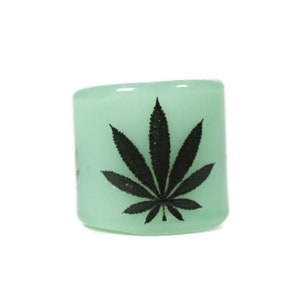 May include: A light green glass ring with a black cannabis leaf design. The ring is cylindrical and has a smooth surface. The cannabis leaf is centered on the ring.
