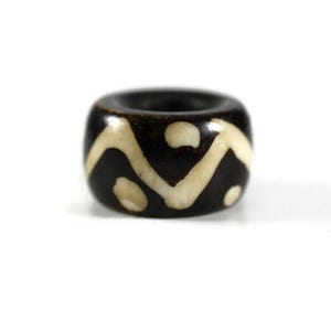 May include: A dark brown, cylindrical bead with a cream-colored, wave-like pattern. The bead has a hole running through the center. The bead is likely made of stone or a similar material. This bead is suitable for jewelry making.