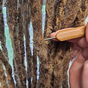 Peut inclure: Gros plan sur des dreadlocks bruns en cours de coiffage avec un crochet à manche en bois. Le crochet est inséré dans la tresse pour la resserrer et maintenir la coiffure. L'arrière-plan est flou, suggérant un cadre intérieur.