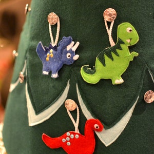 May include: A green felt Christmas tree with three felt dinosaur ornaments. The ornaments are a red dinosaur, a green dinosaur, and a blue dinosaur. The tree is decorated with silver buttons.