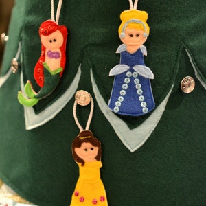 Op de afbeelding: Een groene vilt kerstboom met drie vilt ornamenten. De ornamenten zijn van Ariel uit De Kleine Zeemeermin, Belle uit Belle en het Beest, en Assepoester. Elk ornament is bevestigd aan de boom met een zilveren knoop en touw.