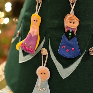 May include: Close-up of a green felt Christmas tree skirt decorated with three felt doll ornaments. The ornaments are in the shape of cartoon characters, with yellow, purple, and blue dresses. Each ornament is attached with a silver button.