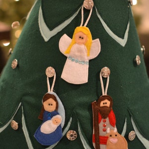 May include: A felt Christmas tree with three felt ornaments: an angel, a woman holding a baby, and a man with a beard and a cow. The ornaments are attached to the tree with silver buttons.