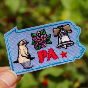 May include: Embroidered patch in the shape of Pennsylvania, featuring a meerkat, pink flowers, and the Liberty Bell. The letters "PA" are in red, with a red star. The patch has a light blue background and a blue border.