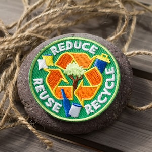 May include: A green, orange, and white embroidered patch with the words "Reduce, Reuse, Recycle" and a stylized recycling symbol with a tree in the center. The patch is attached to a round, dark gray stone.
