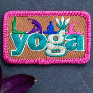 May include: A brown fabric patch with pink stitching around the edges. The patch features the word "yoga" in colorful embroidered letters with various yoga poses in the background.