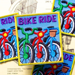 May include: Embroidered patches with a bicycle theme. Each square patch features a colorful bicycle design with the words "BIKE RIDE" in purple lettering on a blue background. The patches are outlined in yellow.