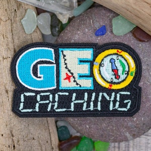 May include: A black and blue embroidered patch with the text "GEO CACHING" and a compass design. The patch is on a rock with sea glass and driftwood.