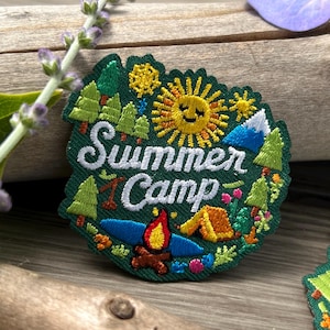 May include: Embroidered patch with a green border featuring a yellow sun, trees, a campfire, and a tent. The words "Summer Camp" are embroidered in white. The patch is designed for outdoor enthusiasts and campers.