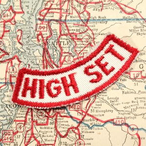 May include: A white patch with red embroidered lettering that reads "HIGH SET" against a vintage map background.