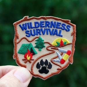 May include: Embroidered patch with the words "WILDERNESS SURVIVAL" in blue. The patch features a tent, campfire, pine tree, and paw print. The patch is tan with brown and blue accents.