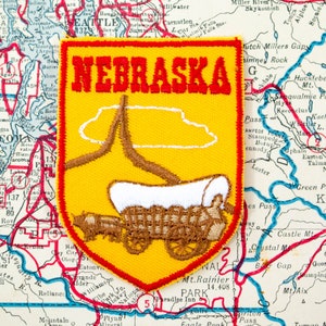 May include: A yellow and red embroidered patch featuring the word "NEBRASKA" in red letters. The patch depicts a covered wagon and a landscape scene, set against a map background.