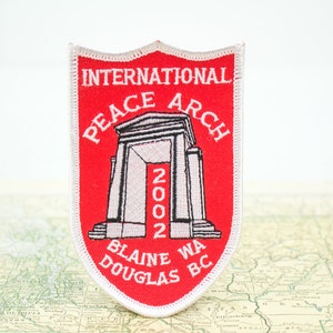 May include: A red and white embroidered patch featuring the International Peace Arch. The patch includes the text "INTERNATIONAL PEACE ARCH 2002 BLAINE WA DOUGLAS BC" in white lettering. The patch is shield-shaped.