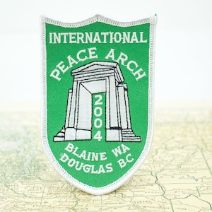 May include: A green and white embroidered patch featuring the International Peace Arch. The patch is shield-shaped and includes the text "INTERNATIONAL PEACE ARCH 2004 BLAINE WA DOUGLAS BC". The patch is placed on a map.