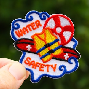 May include: A white embroidered patch with blue, red, and yellow details. The patch depicts a life preserver, a life vest, and a life ring. The text "WATER SAFETY" is embroidered in blue on the patch.