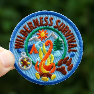 May include: A blue and brown embroidered patch with the text "Wilderness Survival" around the edge. The center of the patch features a campfire with a compass, tent, mountains, and a pine tree.
