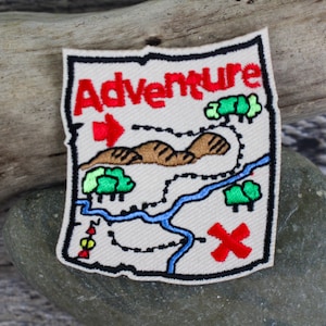 May include: Embroidered patch with the word "Adventure" in red, featuring a map design with a red arrow, brown mountains, green trees, a blue river, and a red "X" marking a destination. The patch has a cream-colored background and a black border.