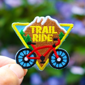 May include: A triangular embroidered patch with a mountain scene and a red bicycle. The text "TRAIL RIDE" is embroidered in yellow on the patch.