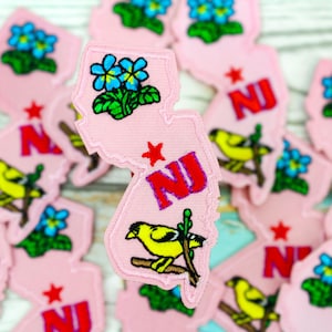 May include: A collection of pink felt patches shaped like the state of New Jersey. Each patch features a different design, including a yellow bird, blue forget-me-not flowers, and the letters "NJ" in red and blue.